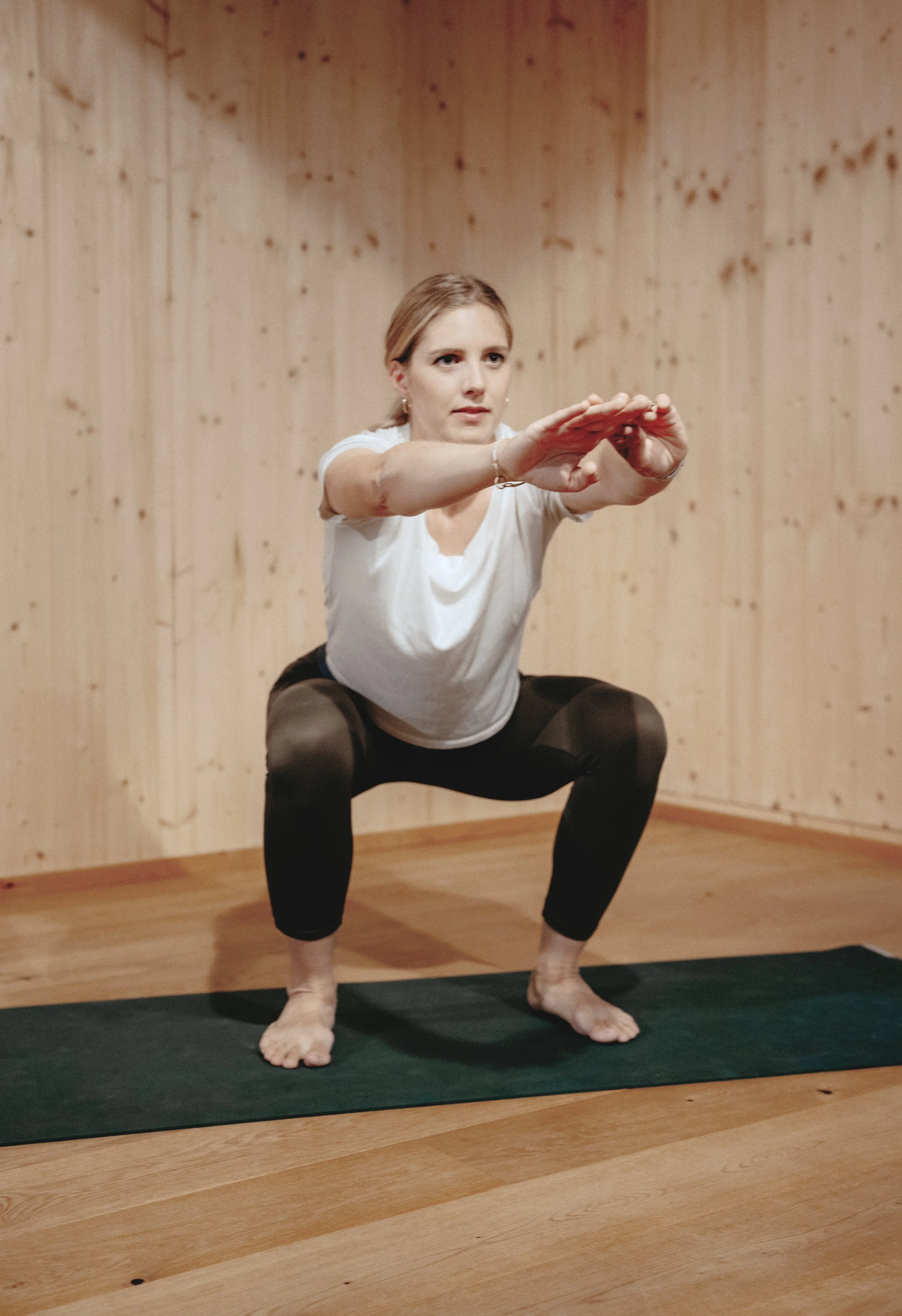 Eine Frau macht eine Squat-Übung auf einer Yogamatte in einem Raum mit Holzwänden.