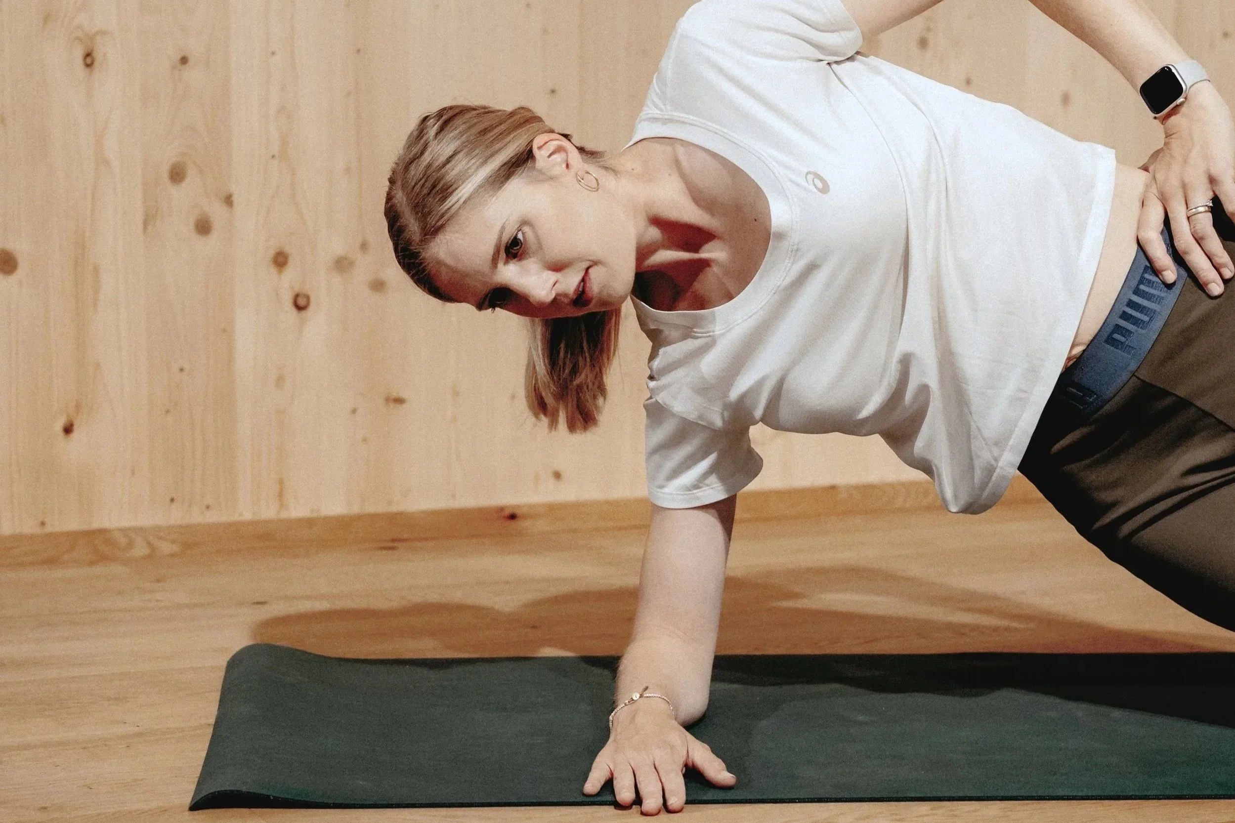 Frau beim Yoga auf einer schwarzen Matte, die auf einem Holzfußboden vor einer Holzpaneelwand sitzt, mit ausgestrecktem Arm nach oben, im Crow Pose.