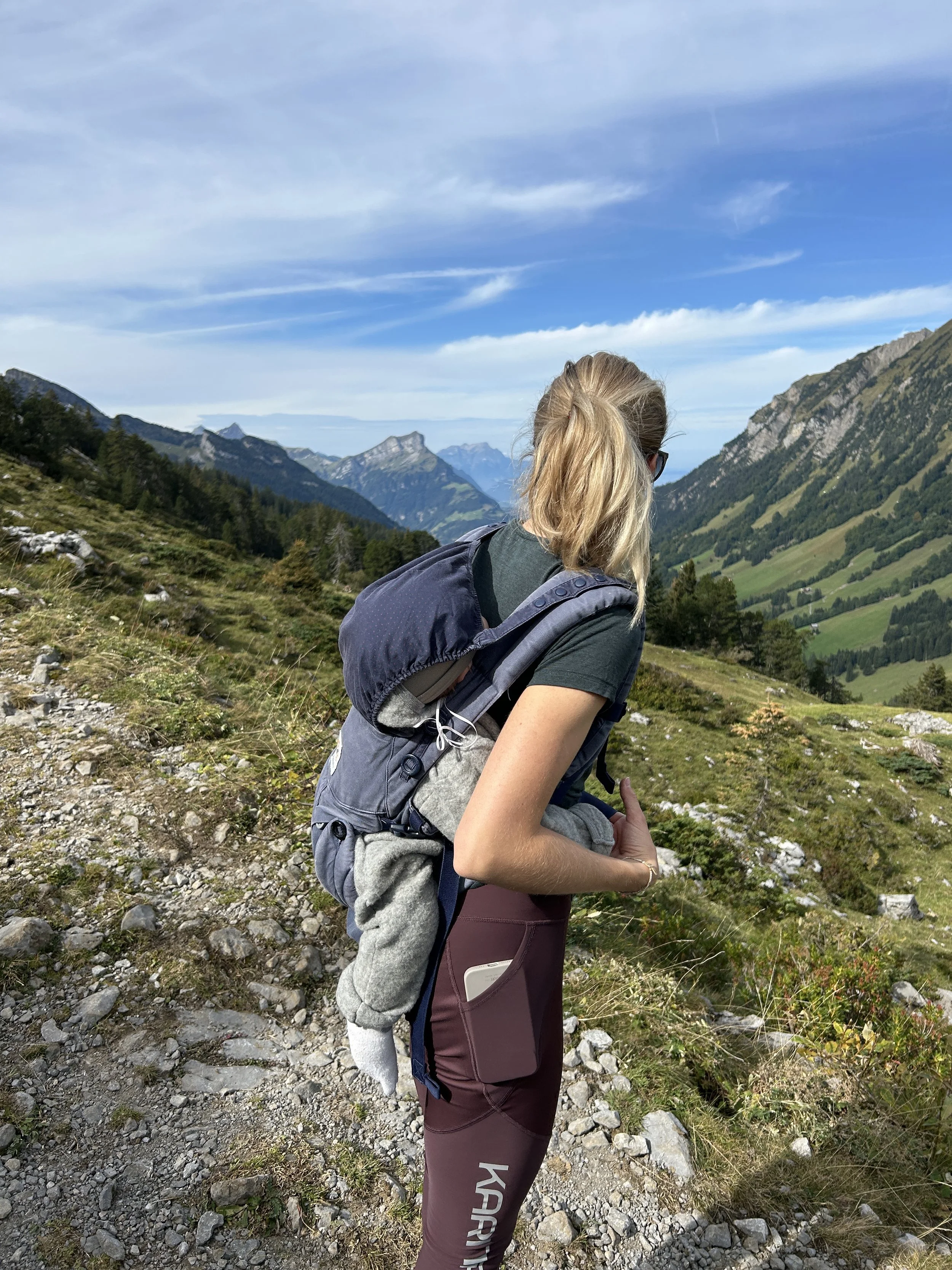Eine Frau mit blondem Haar in einem grauen T-Shirt und lila Hosen, die einen Rucksack trägt, steht auf einem Bergpfad in den Bergen, schaut auf die Landschaft mit bewaldeten Hügeln und Bergen im Hintergrund, bei schönem Wetter mit blauen Himmel und einigen Wolken.