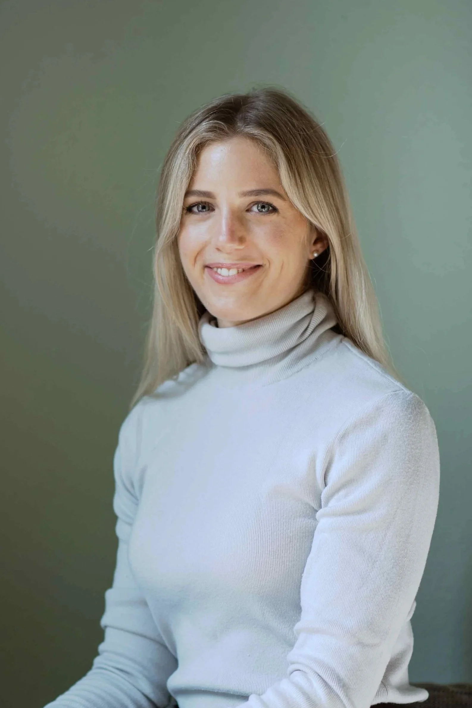 Porträt einer lachenden Frau mit blonden, schulterlangen Haaren, blauen Augen und hellhäutiger Haut, die ein weißes Turtleneck-Shirt trägt, vor einem grünen Hintergrund.