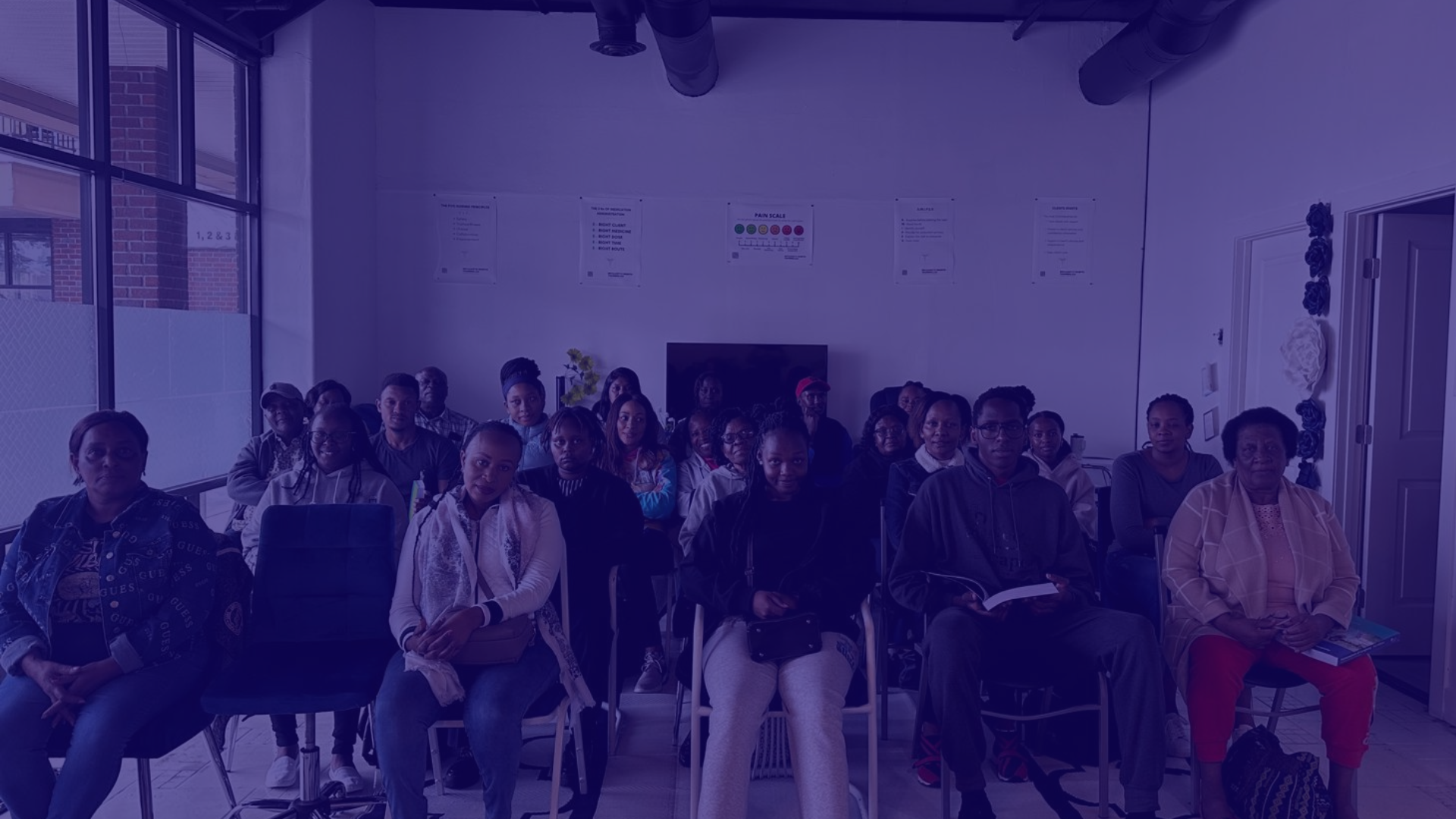 Group of diverse adults seated in a room, attending a presentation or meeting, with posters on the wall and large windows letting in natural light.