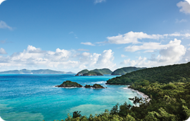 Tropical ocean coastline with turquoise waters, rocky outcrops, lush green hills, and a partly cloudy sky.