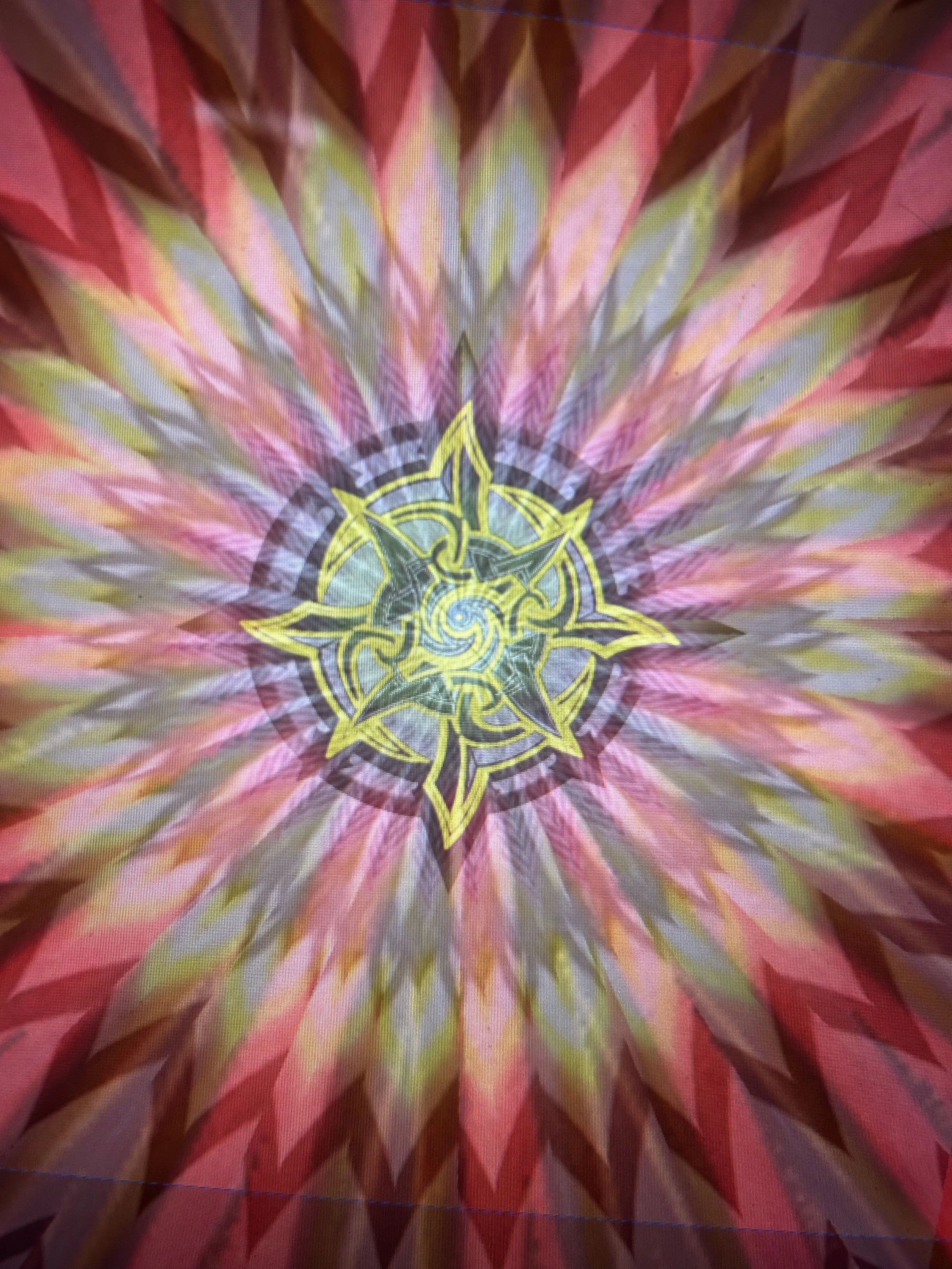 Abstract kaleidoscopic pattern with vibrant pink, yellow, green, and purple colors forming a starburst design with a central geometric motif in yellow and black.