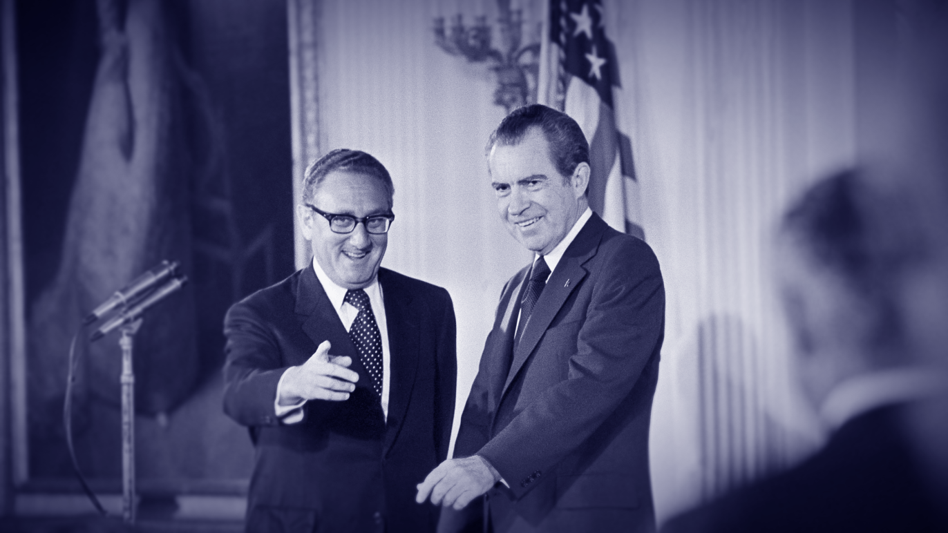 Two men in suits smiling and shaking hands during a formal event, with an American flag and a flag with stars and stripes in the background, and a microphone on a stand nearby.