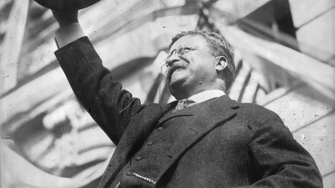 A black-and-white photo of a man with a mustache and glasses, smiling and raising his arm in victory or celebration.