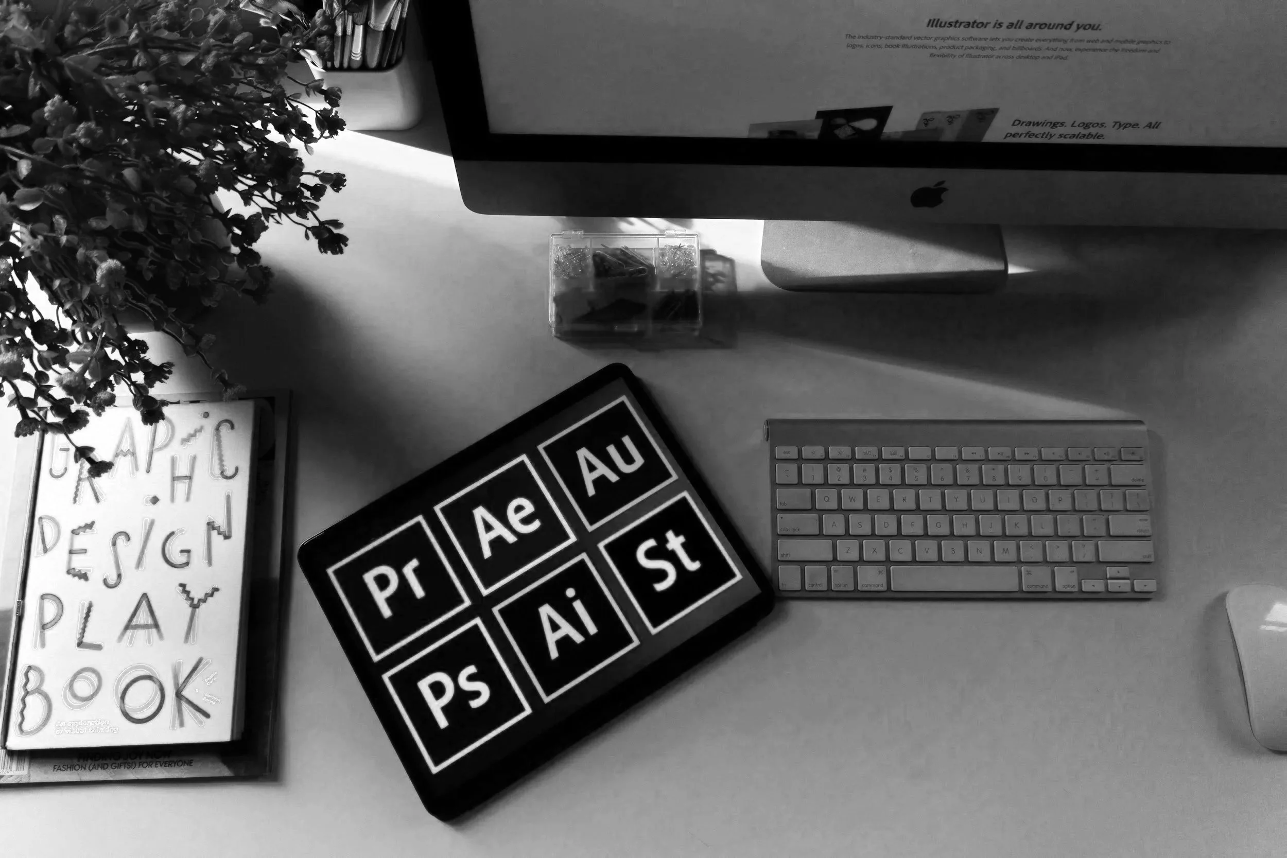 Black and white photo of a desk with computer monitor, keyboard, and stationery, including a jar of colored pencils. There are fashion and typography books and design stickers on the desk, along with a potted plant on the left side.