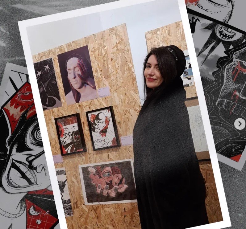 Young woman with black hair, wearing a black coat and hat, standing beside an art display on a wooden board with various artworks.