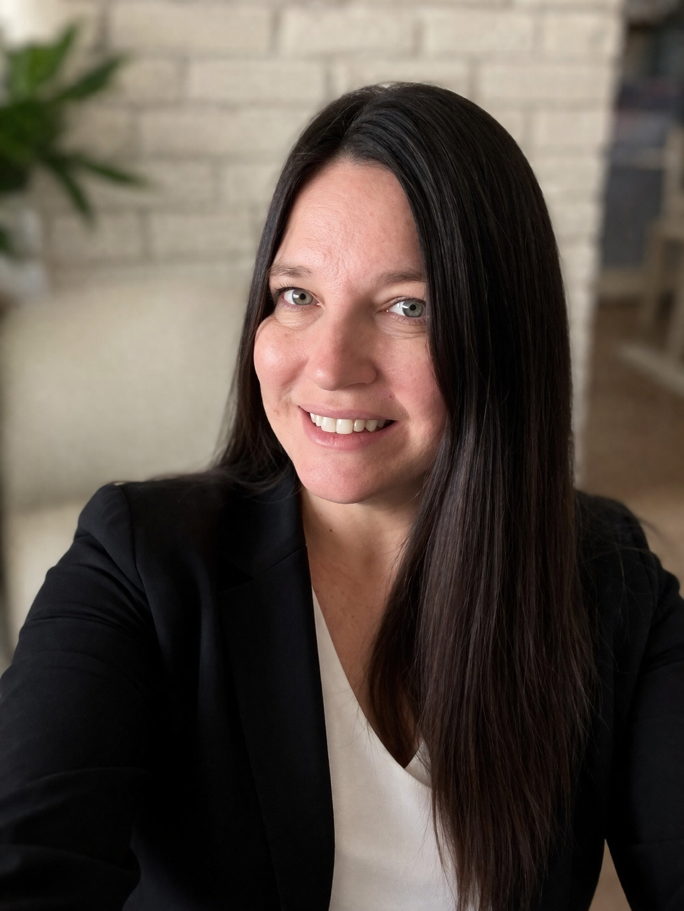 A woman with long dark hair smiling in a black blazer and white top, with a blurred brick wall background and some greenery.