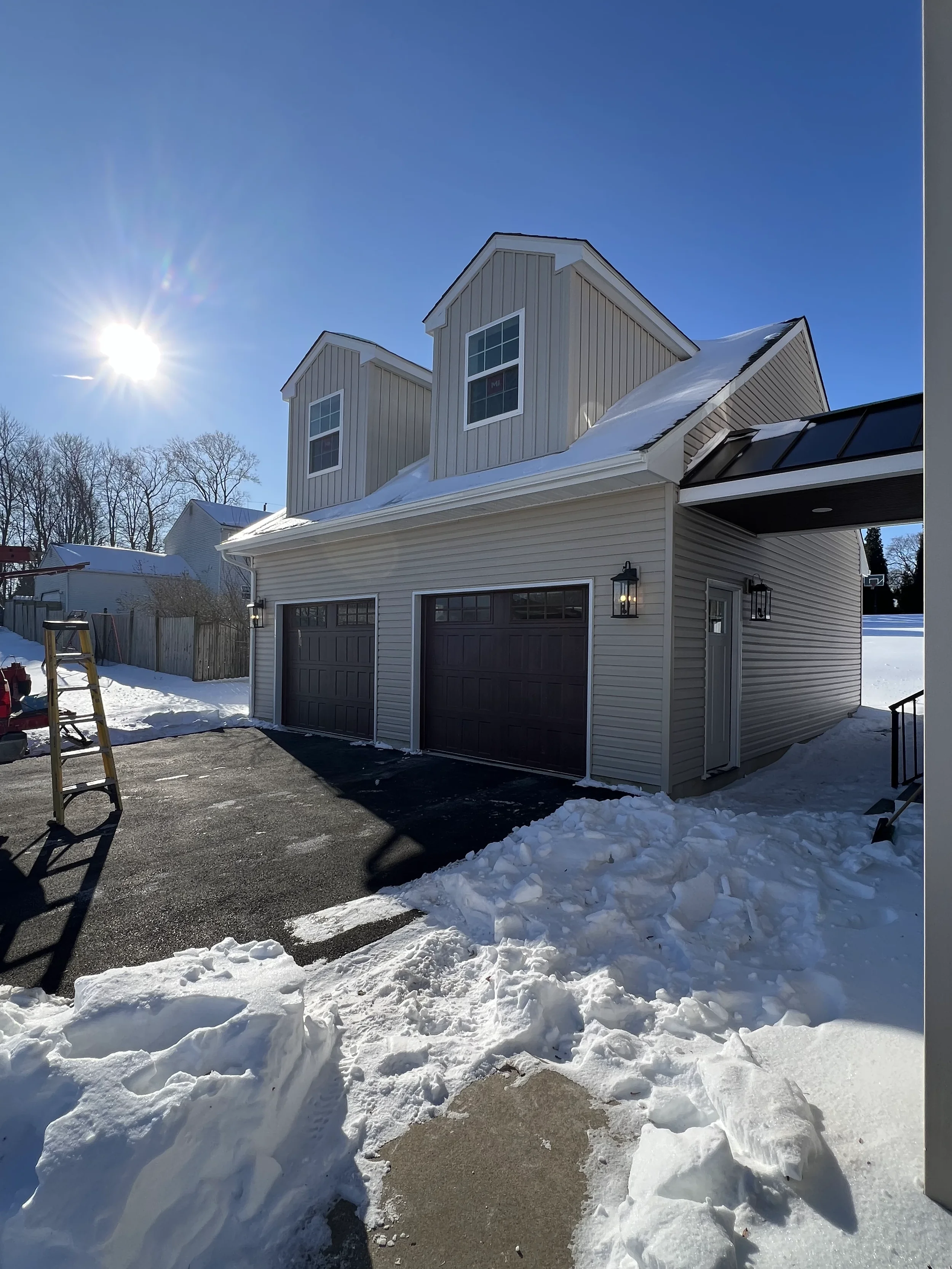 A modern house with beige siding and a garage with black doors, surrounded by snow, under a clear blue sky with the sun shining brightly.