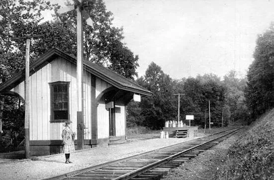 Black and white photo of a small train station with a woman standing on the platform. Railroad tracks run alongside the station, with trees and utility poles in the background.