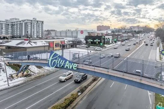 A city street scene with a pedestrian bridge labeled 'City Ave,' snow-covered ground, cars driving on the road, commercial buildings including Target, and apartment complexes in the background, under a partly cloudy sky during sunset.