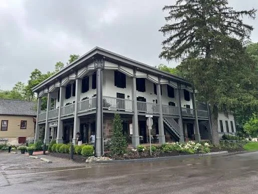 A two-story building with a covered porch and stairs, surrounded by bushes and trees, on a cloudy day.