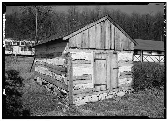 A black-and-white photo of a small wooden shed with a sloped roof, situated on a grassy area with trees in the background.
