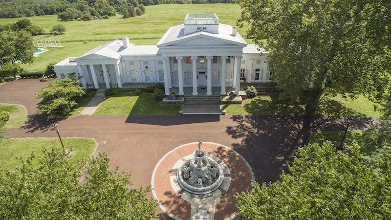A large white mansion with a portico and columns, surrounded by greenery. There is a circular fountain in front of the entrance and a brick driveway leading to the house.