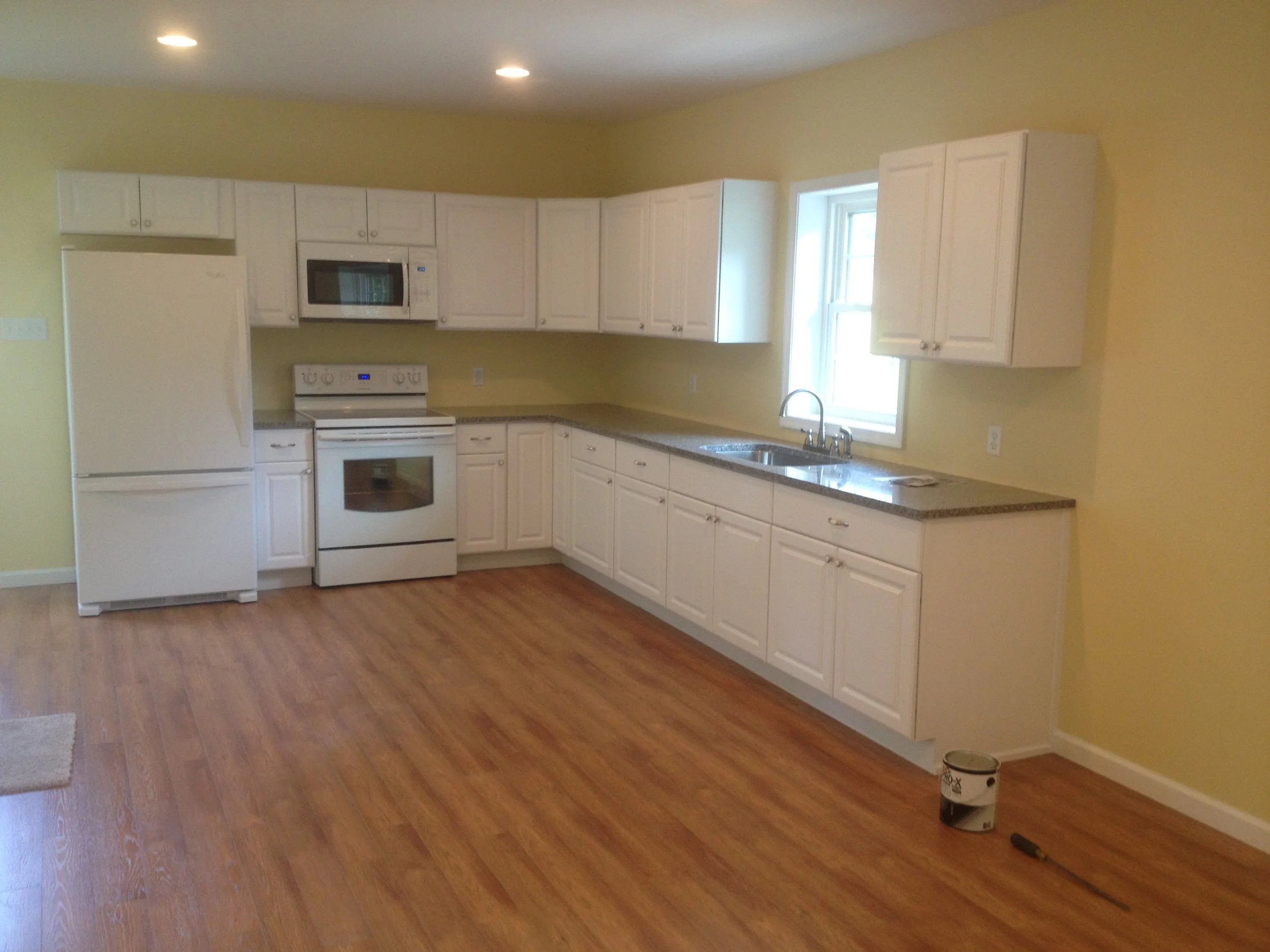 Kitchen with white cabinets, yellow walls, wooden flooring, and a large window above the sink. There is a stove, microwave, refrigerator, and a can of paint on the floor.
