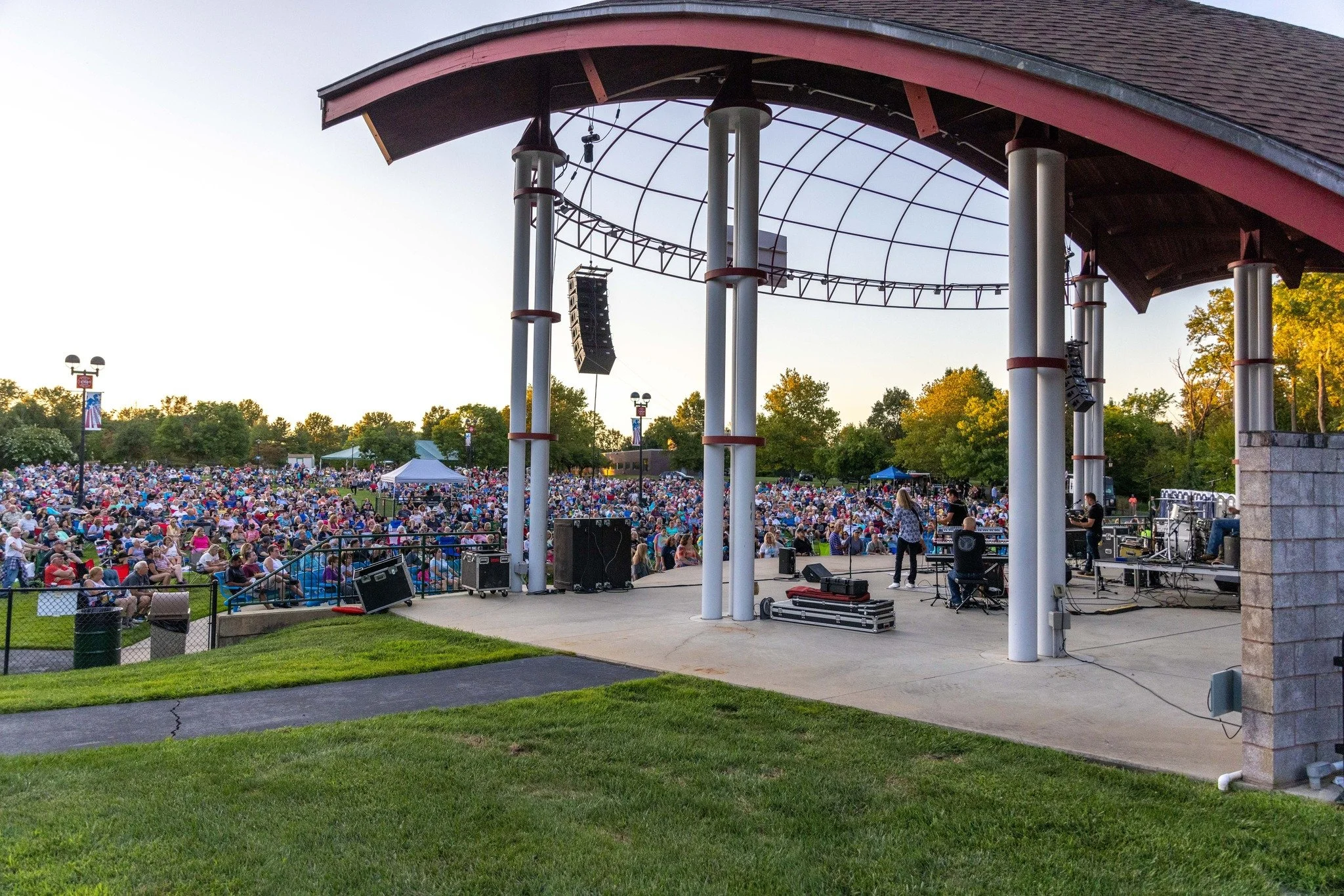 Outdoor concert stage with a crowd gathering in a park during sunset, featuring a band setup with instruments and speakers under a pavilion.