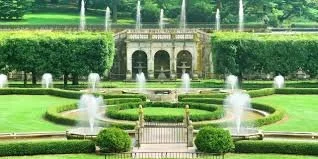 A landscaped garden with symmetrical water fountains, green bushes, and a stone structure in the background.