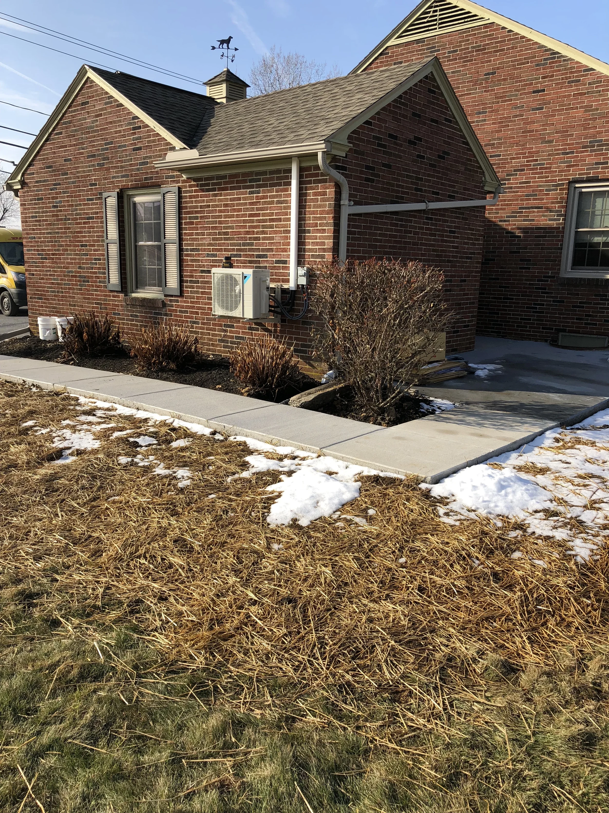 A brick house with a small yard, some trimmed bushes, a sidewalk, patches of snow, and a clear blue sky.