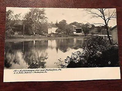 A vintage black-and-white photo of a river with trees and houses in the background.