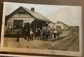 Black and white photo of a vintage train station with a group of people standing on the platform, some holding suitcases, and a train visible on the right side.