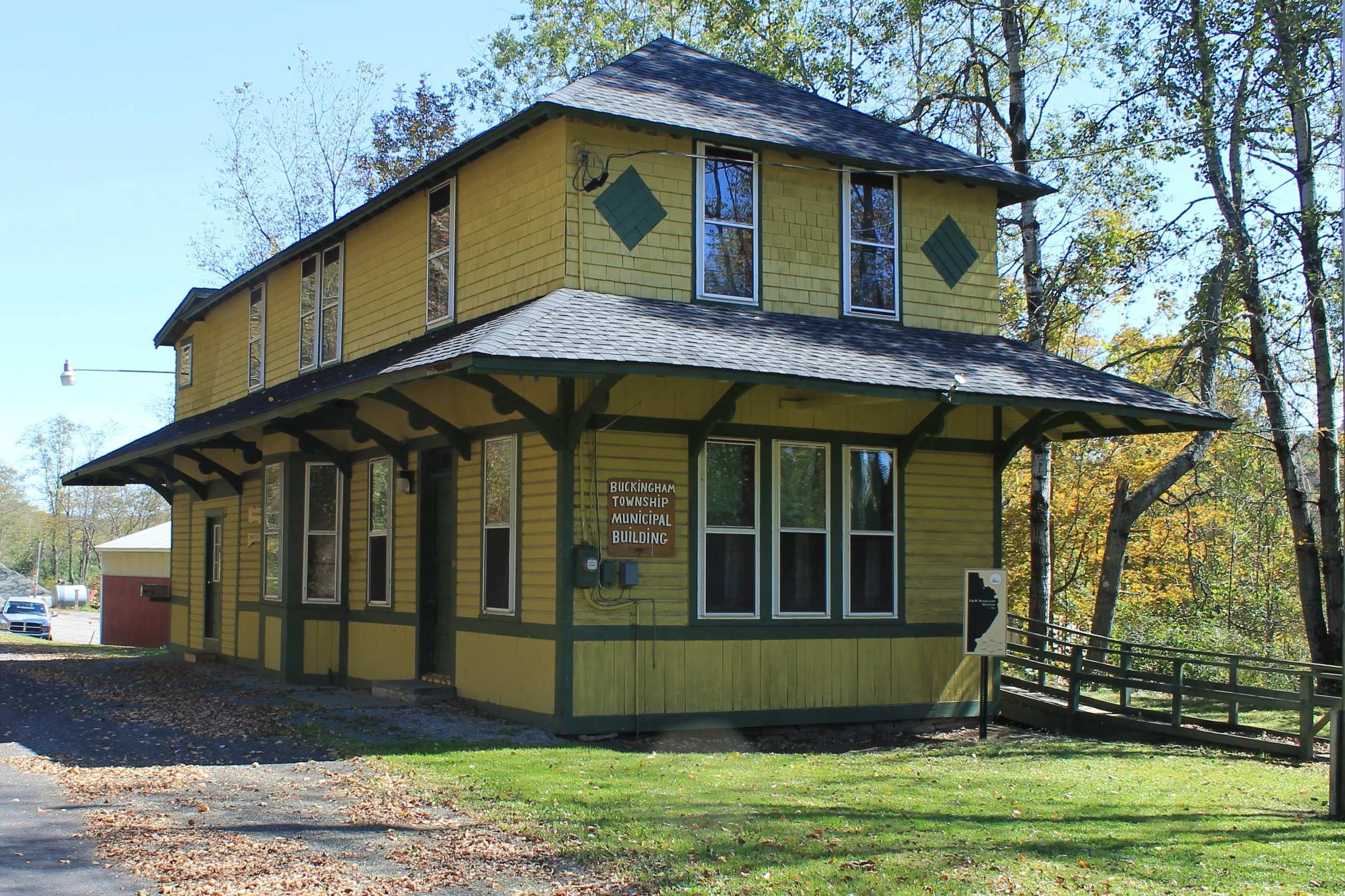 Yellow wooden building with multiple large windows and a sign that reads 'Buckingham Township Municipal Building', surrounded by trees with autumn foliage.