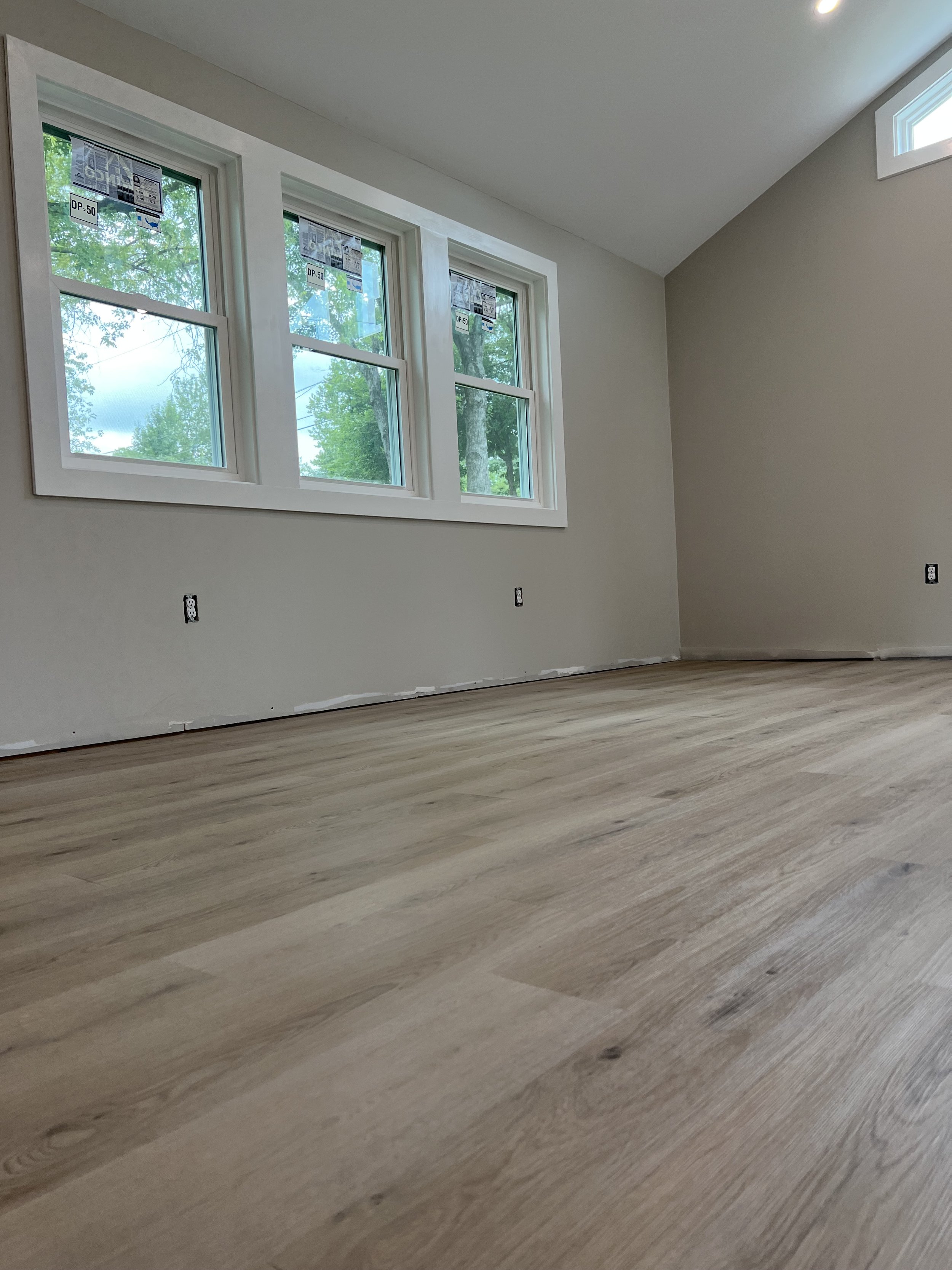 Interior of a room under construction with three large windows, light-colored walls, and unfinished electrical outlets, showing a wood floor and green trees outside.