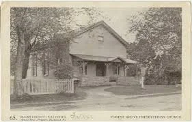 A vintage black and white photo of a two-story wooden house with a porch, picket fence, and trees in the yard.