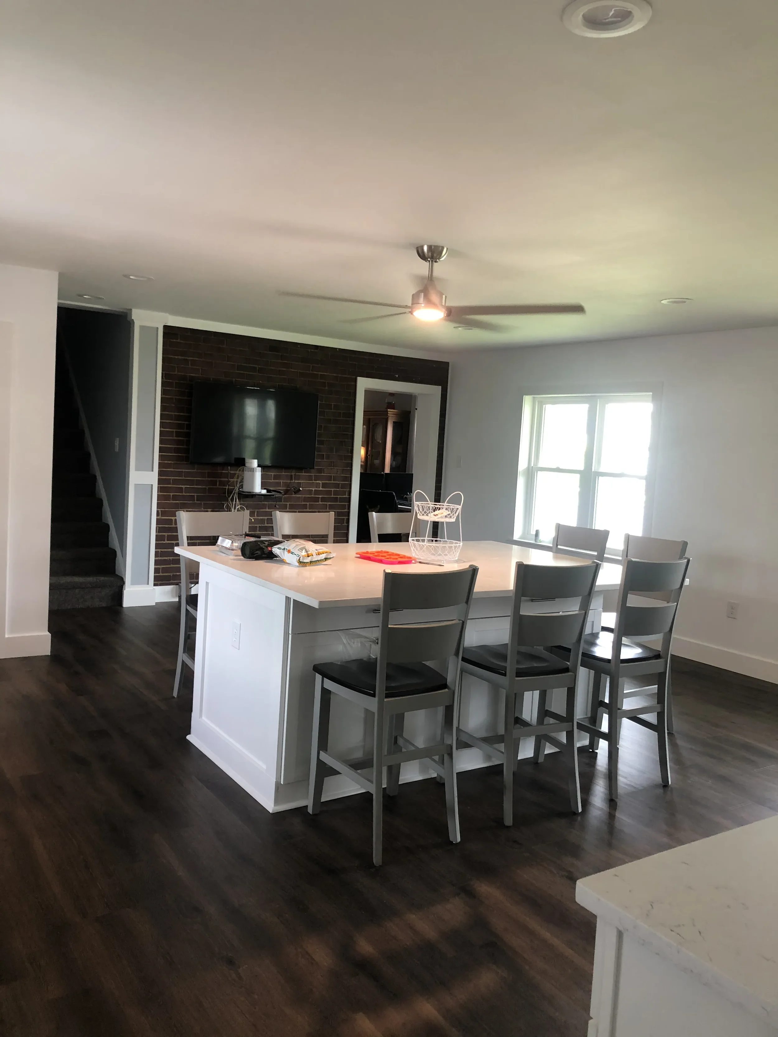 A dining area with a white island table, six chairs, a brick accent wall, a wall-mounted television, and a ceiling fan with lights. There are windows on the right side and a staircase to the left.