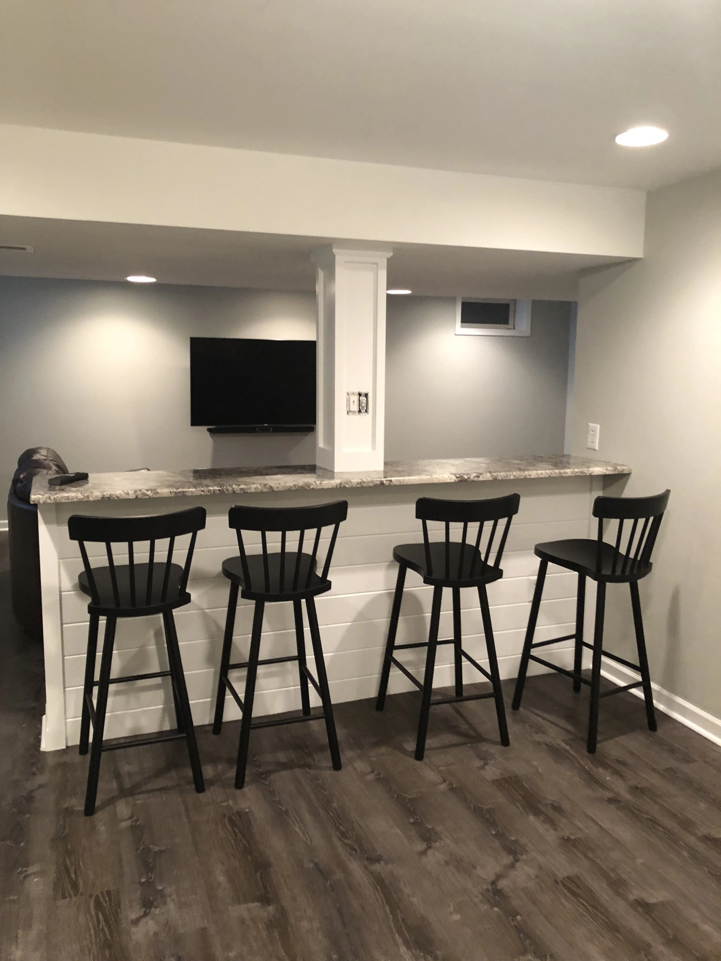 View of a modern kitchen island with black chairs, granite countertop, and a mounted flat-screen TV on the wall in the background. Recessed lighting in the ceiling and a small window.