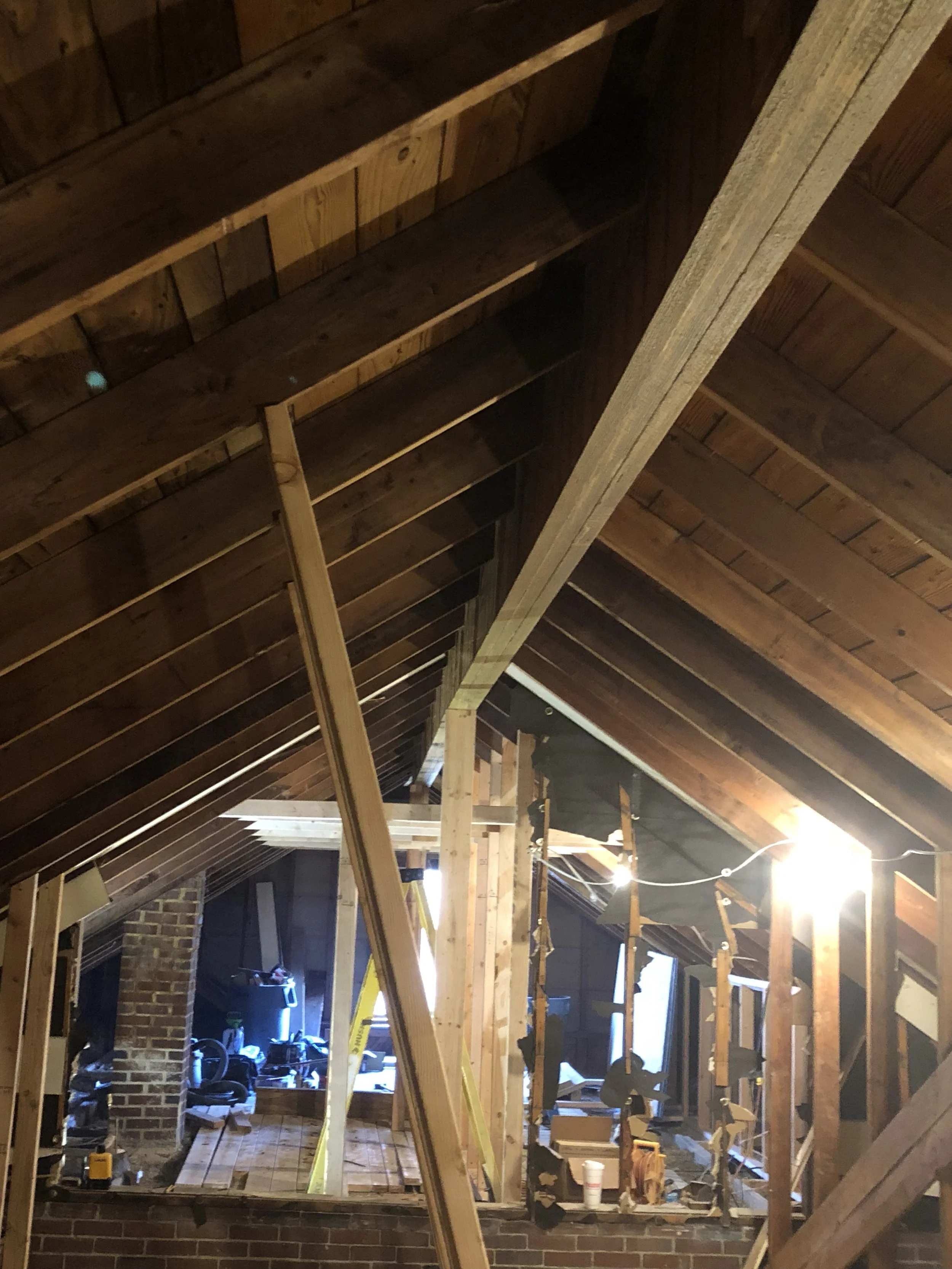 View of a partially constructed attic space with exposed wooden beams and framing, scattered construction tools and materials, and a ladder, under warm lighting.