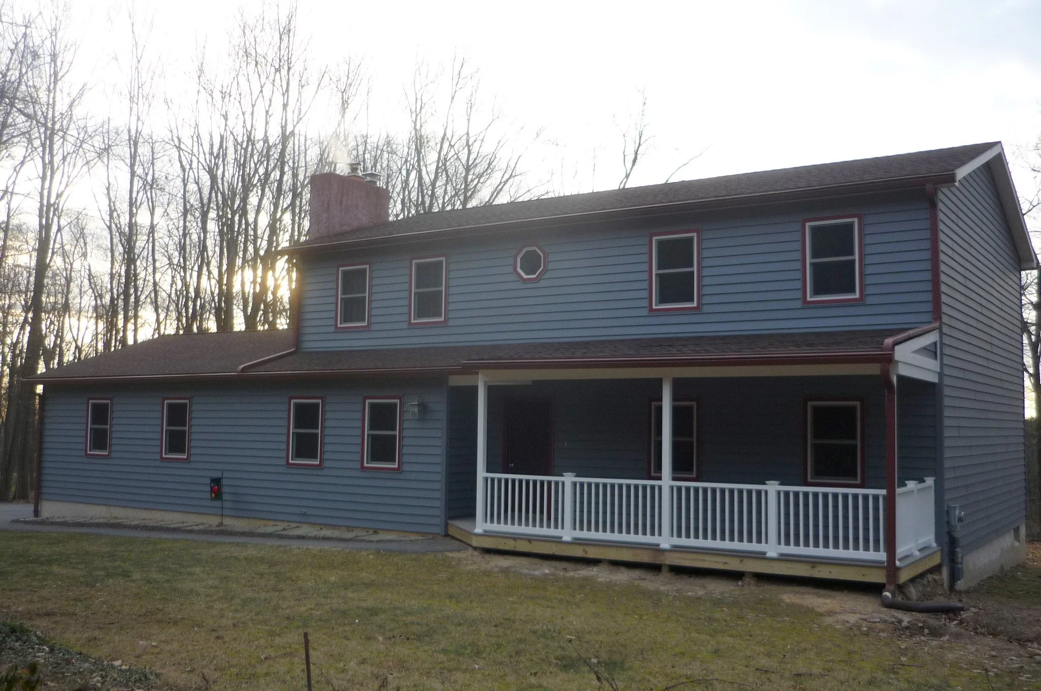 A two-story house painted blue with red trim around the windows, a white porch railing, and a brown roof, surrounded by leafless trees with the sun setting behind.