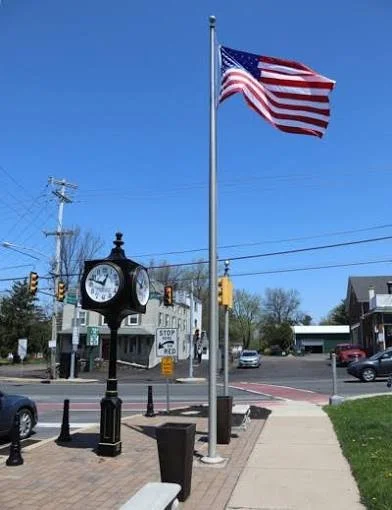 American flag flying at half-staff next to a clock on a small town sidewalk with traffic lights and a building in the background.