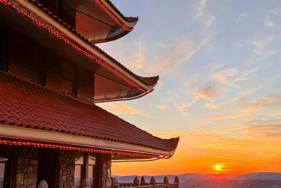 A traditional Asian-style building with curved red-tile roofs against a sunset sky, with a mountain range in the background.