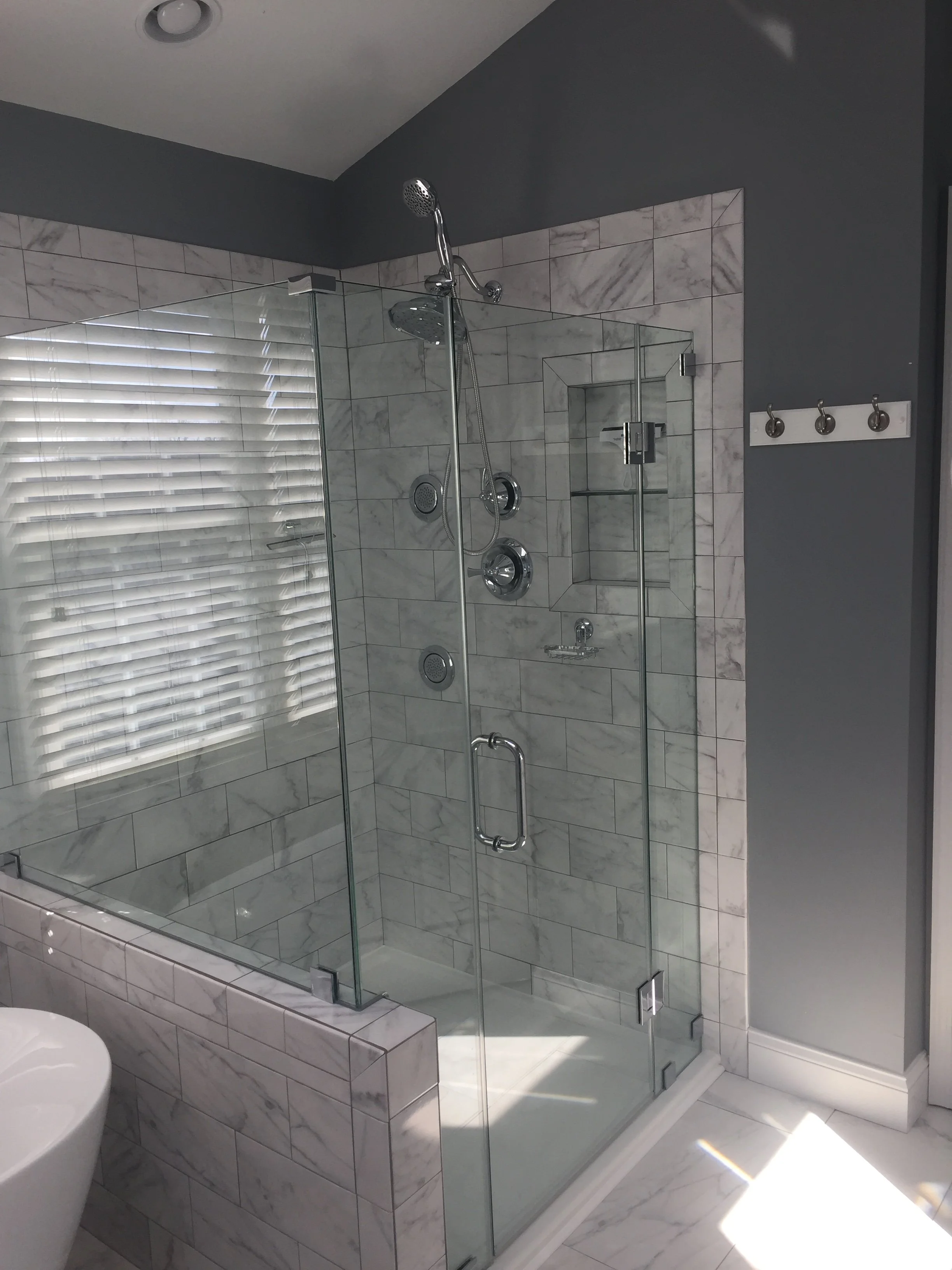 Modern walk-in shower with glass enclosure, marble tiles, a built-in shelf, showerhead, and multiple shower controls, in a bathroom with gray walls and a window with blinds.