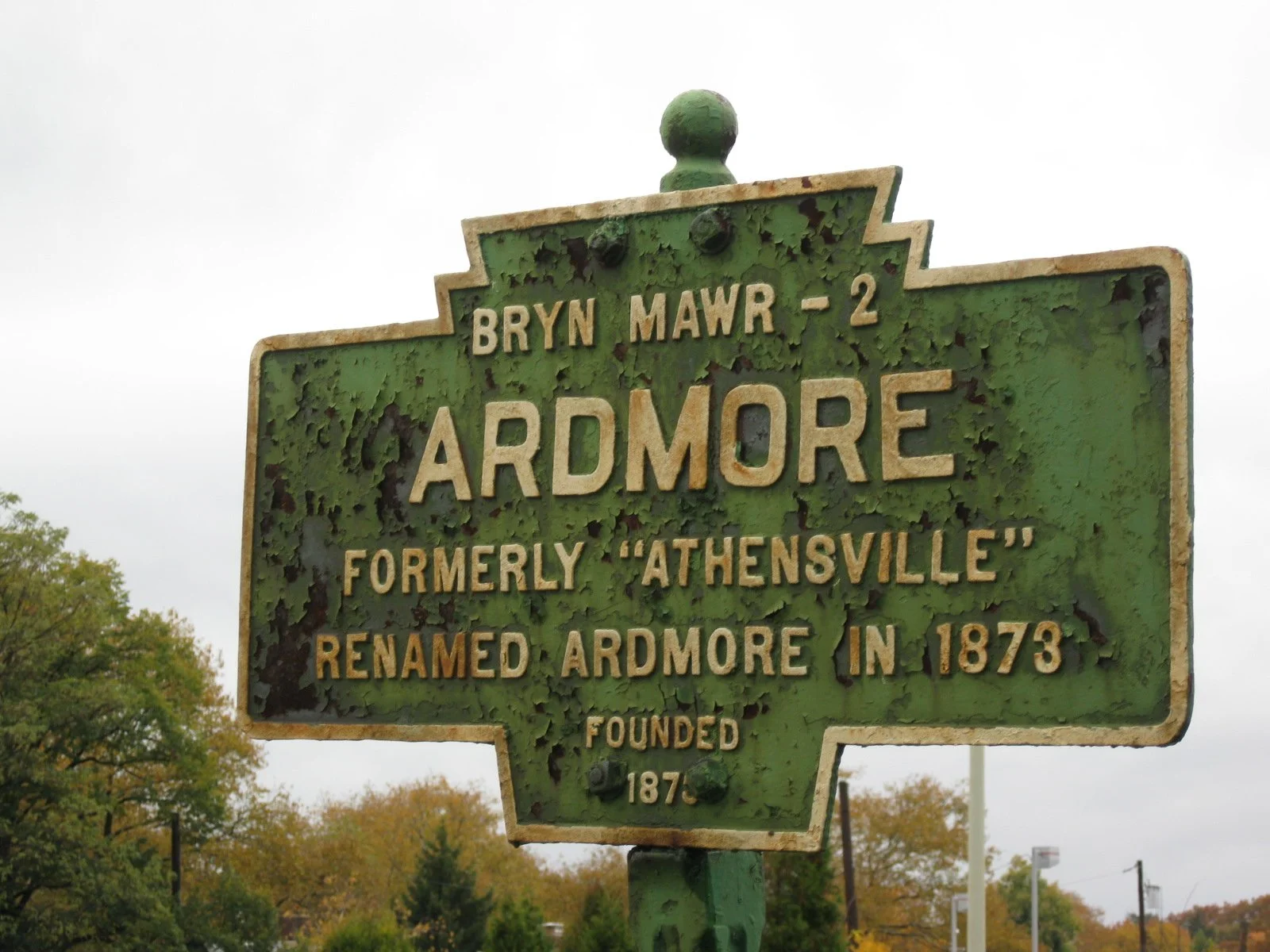 Historical green street sign for Ardmore, formerly Athensville, indicating it was renamed in 1873, with visible rust and peeling paint, against a cloudy sky and trees in the background.