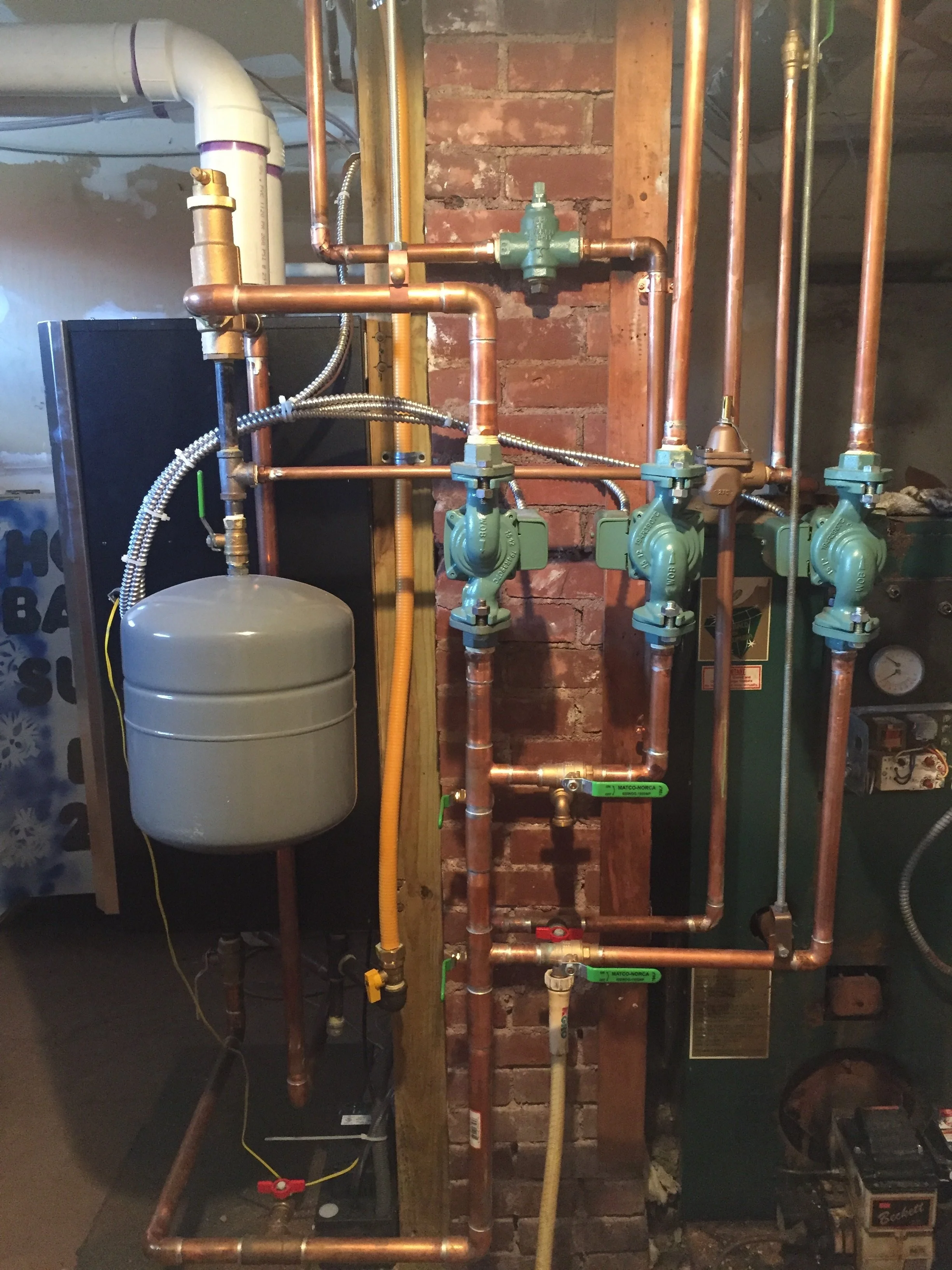 A plumbing system with copper pipes, valves, and a pressure gauge mounted against an unfinished brick wall in a basement or utility room.