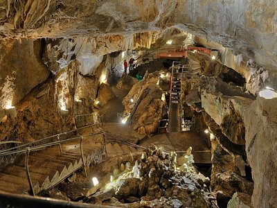 Inside a large cave with rocky walls and stalactites, featuring a walkway with railings and lights, leading deeper into the cave.