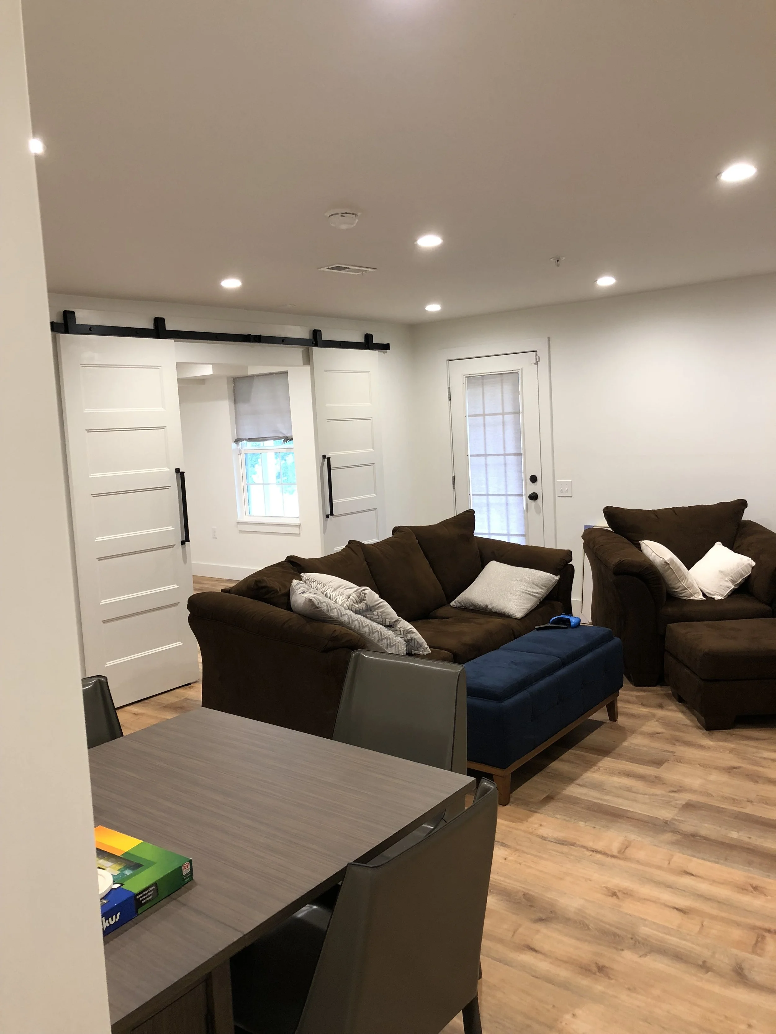 Living room with a brown sectional sofa, white pillows, and a wooden floor, adjacent to a dining table with chairs, against white walls, and illuminated ceiling lights.
