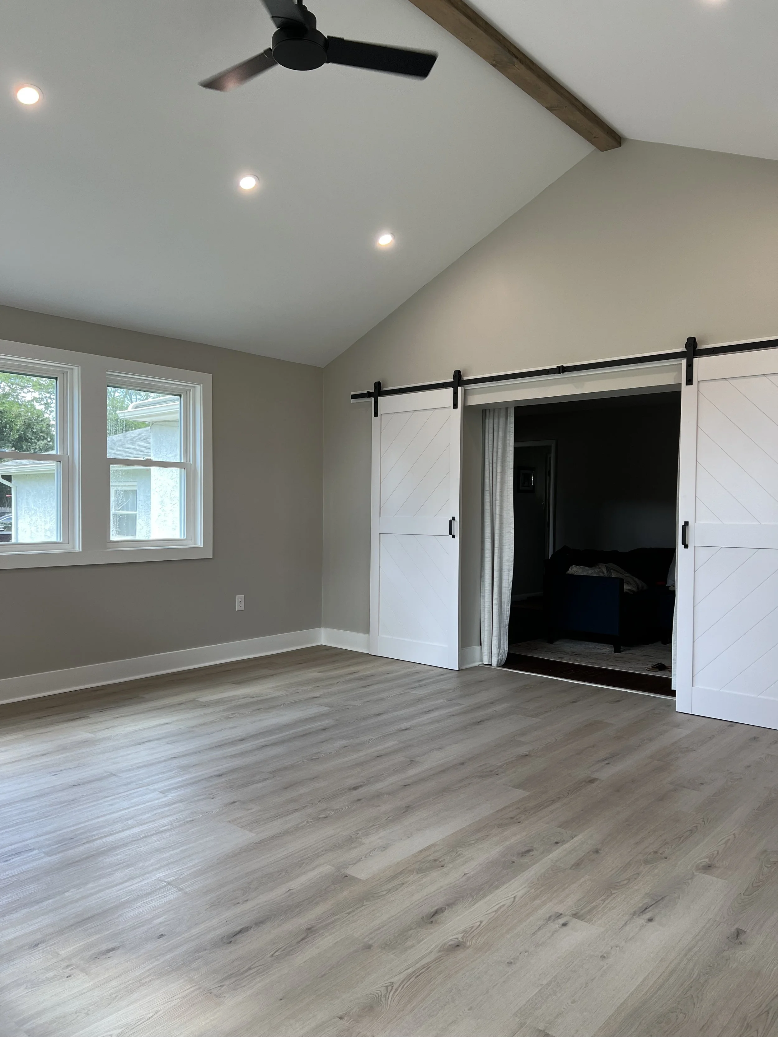 A room with beige walls, large windows, light-colored wooden flooring, a ceiling fan, recessed ceiling lights, and sliding barn doors leading to another room.