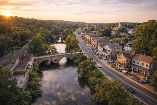 Aerial view of a small town at sunset with a river running through it, old stone bridges, and a main street lined with shops and cars.