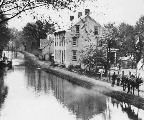 Black and white photo of a large house next to a canal with people and horses on the sidewalk.