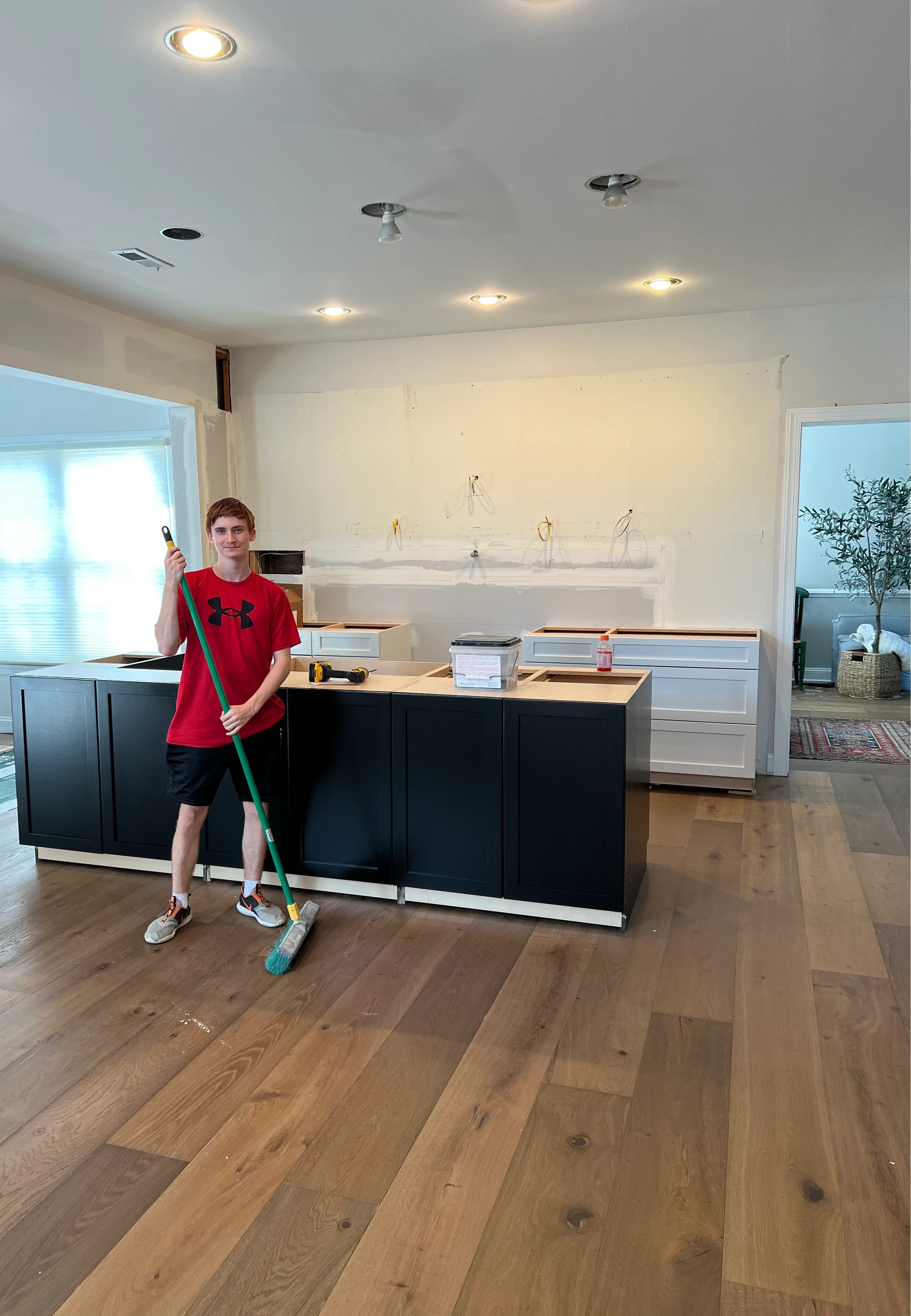 A person with short brown hair wearing a red Under Armour t-shirt, black shorts, and athletic shoes is cleaning a hardwood floor with a mop in a kitchen that is under construction. The kitchen has black cabinets and white drawers, a partially finished white wall, and visible ceiling lights and wiring.