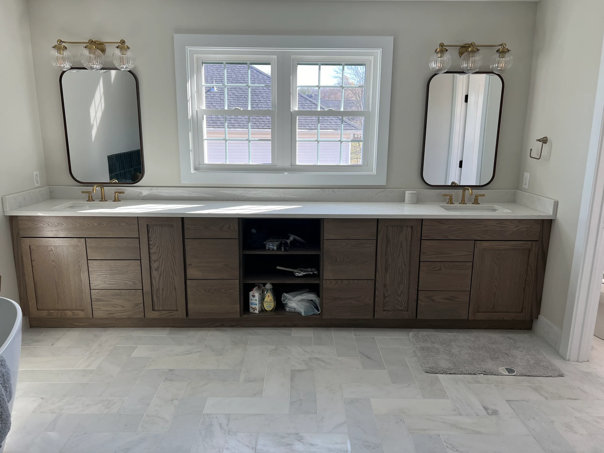 Bathroom vanity with two mirrors, brass faucets, window above, and various cleaning supplies and items below, with beige tiles on the floor.
