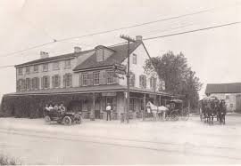 A black-and-white photo of a multi-story building with a porch, with vintage cars parked in front and horse-drawn carriages nearby.