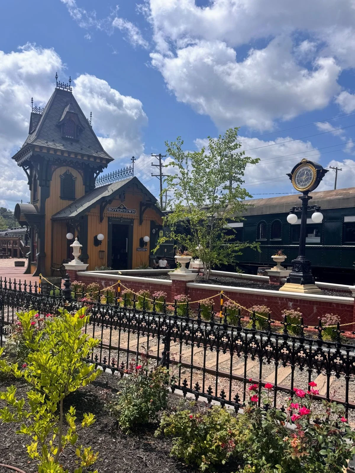 Boyertown historic train station with a vintage clock, a black train car, and a garden with plants and flowers under a partly cloudy sky.