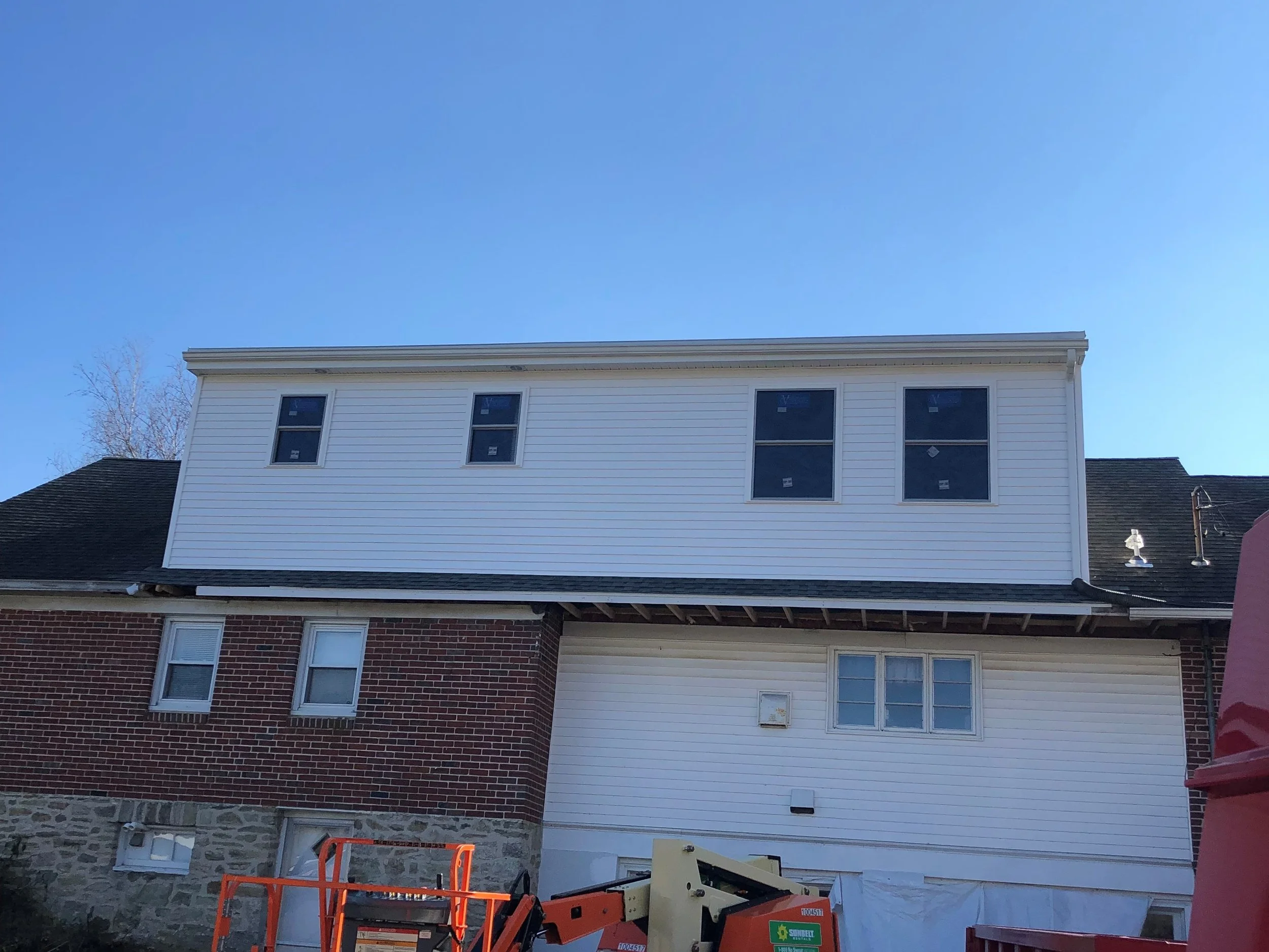 The photo shows the upper part of a multi-story house with white siding and several windows. Below, part of the roof is visible with a stone facade at the basement level. Construction equipment, including an orange lift and some tools, are in the foreground. The sky is clear and blue.