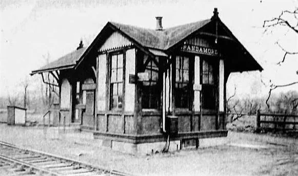 Old black and white train station building with a sign that reads 'Randall' and train tracks in front.
