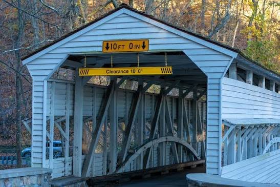 The image shows a covered bridge with a blue exterior, with height clearance signs reading 10 feet 0 inches, and black and yellow-striped warning signs, set in a wooded area.