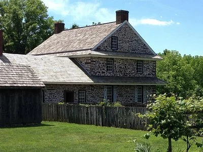 A large stone house with a steep shingled roof, multiple chimneys, and a wooden fence in front. The house has several small windows and is surrounded by green trees and grass.