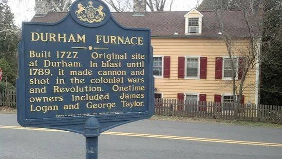 Historical marker for Durham Furnace, built in 1727, located at its original site in Durham. The furnace was used until 1789 to make cannon and shot during colonial wars and the Revolution. Owners included James Logan and George Taylor. In the background, there is a two-story house with a sloped roof, beige siding, and red shutters, along with a wooden fence and trees.